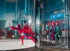 A person in a red suit and helmet is indoor skydiving in a wind tunnel while an instructor and four observers watch from outside, all wearing helmets and jumpsuits.