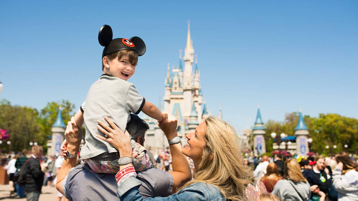 A young boy wearing Mickey Mouse ears sits on his father’s shoulders, with his mother smiling beside them in front of Cinderella’s Castle at Walt Disney World in Orlando, Florida.