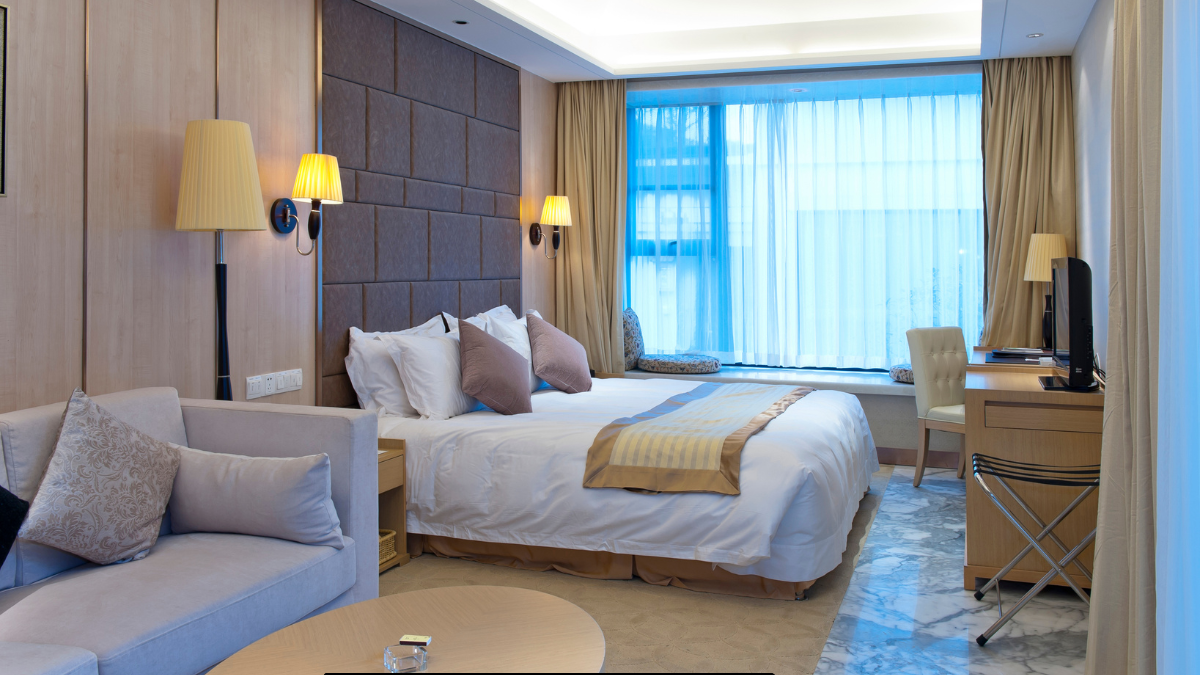 Modern hotel room with a king-size bed, beige decor, wall-mounted lamps, a small sofa, and a desk by a large window with sheer curtains.