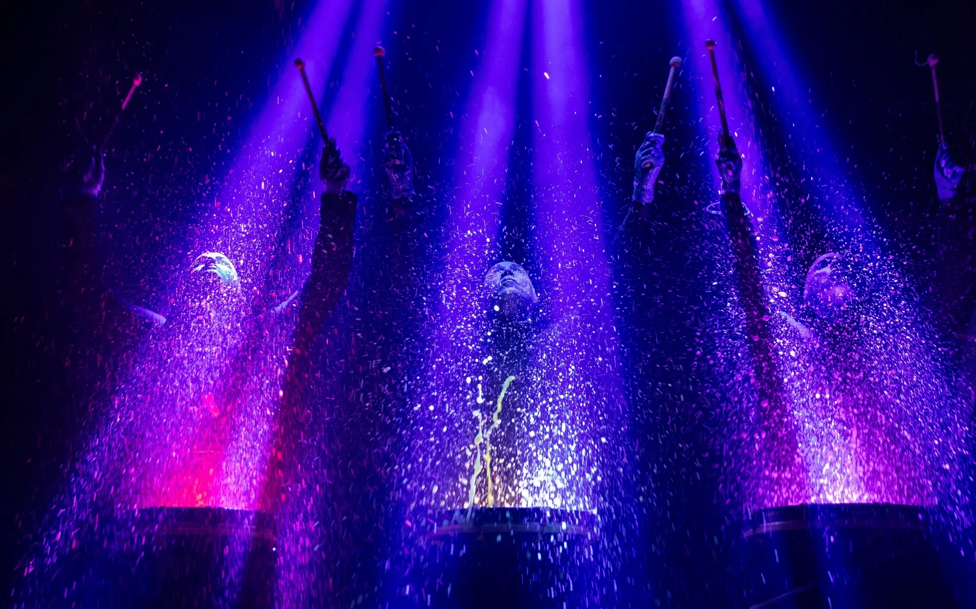 Performers in dark costumes raise drumsticks under vivid purple stage lights as colorful paint or water splashes dramatically around them.