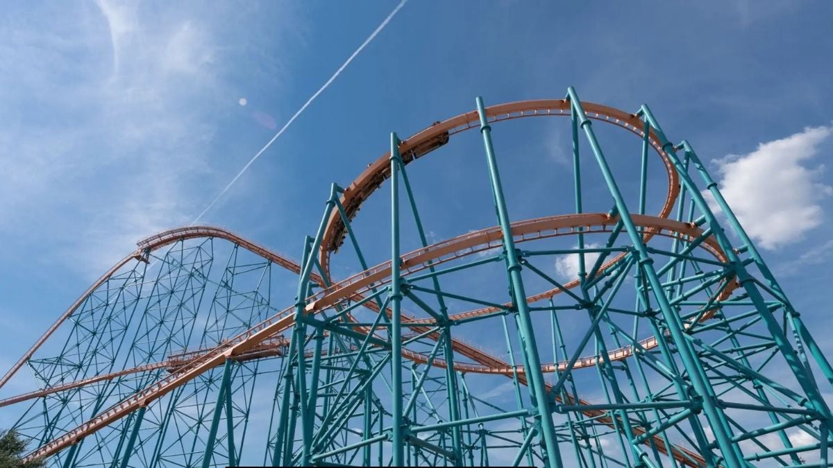 A roller coaster winds through sweeping orange track and teal support beams at Six Flags America in Maryland under a bright blue sky with thin clouds.