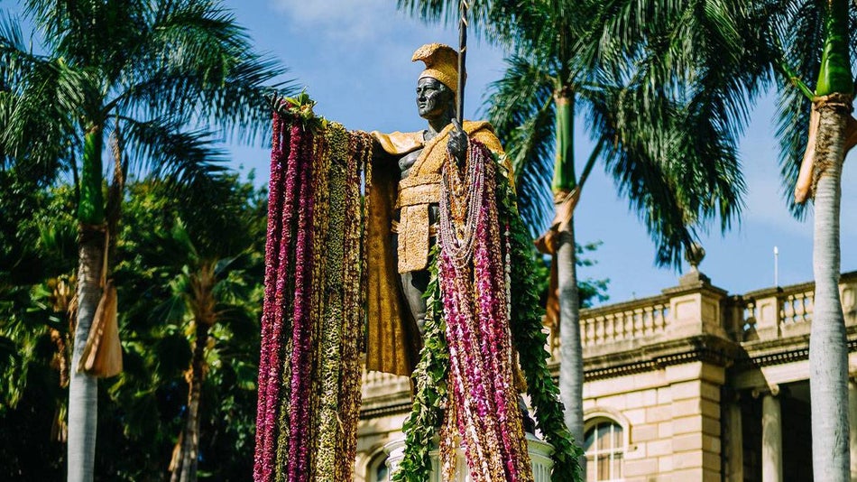 close up of Kamehameha Statue adorned with lays surrounded by palm trees and building in background - The Big Island, Hawaii, USA