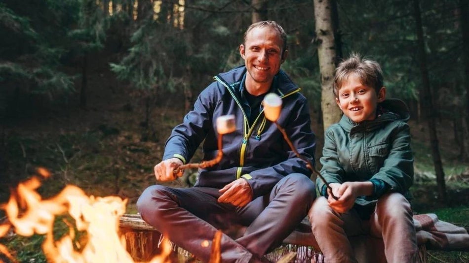 father and son sitting at campfire together in the woods roasting marshmellows