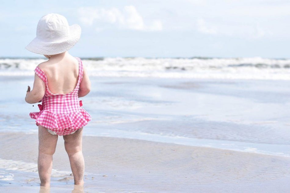 16 month old baby standing on beach with white sunhat and pink gingham swimsuit