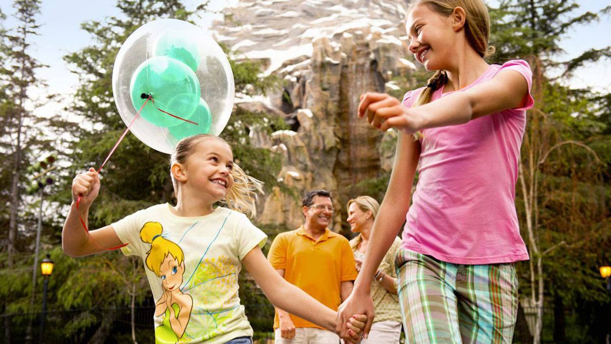 two girls holding hands and skipping with Disney balloons and parents in background at Disneyland in Los Angeles, California, USA