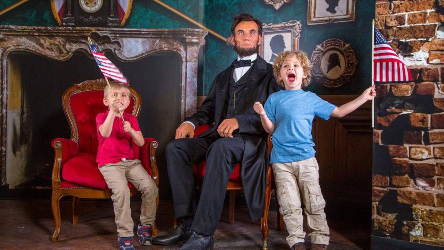 two excited kids holding united states flag sitting next to wax figure Abraham Lincoln at Madame Tussauds in Orlando, Florida, USA