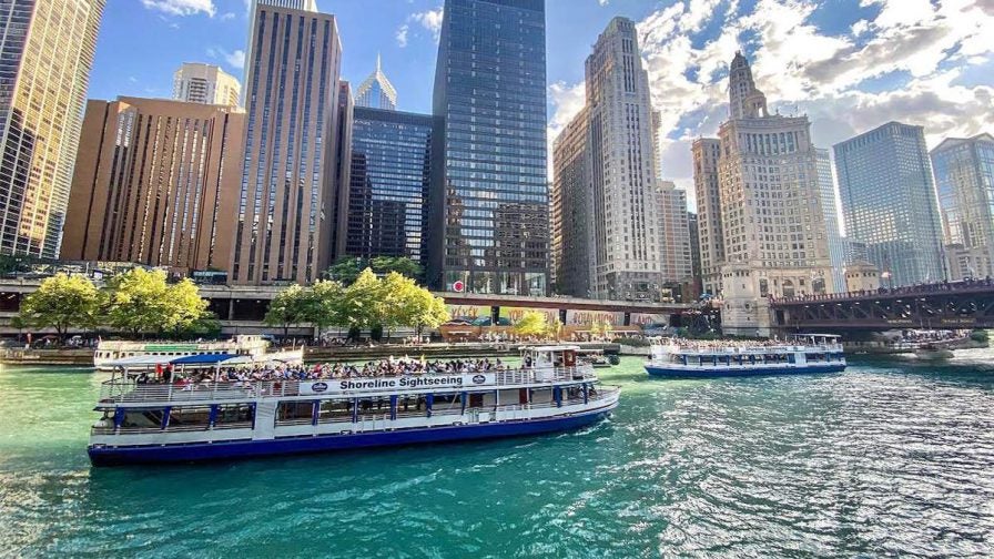 Wide shot of a boat doing a river tour with large city buildings behind it on a sunny day in Chicago, Illinois