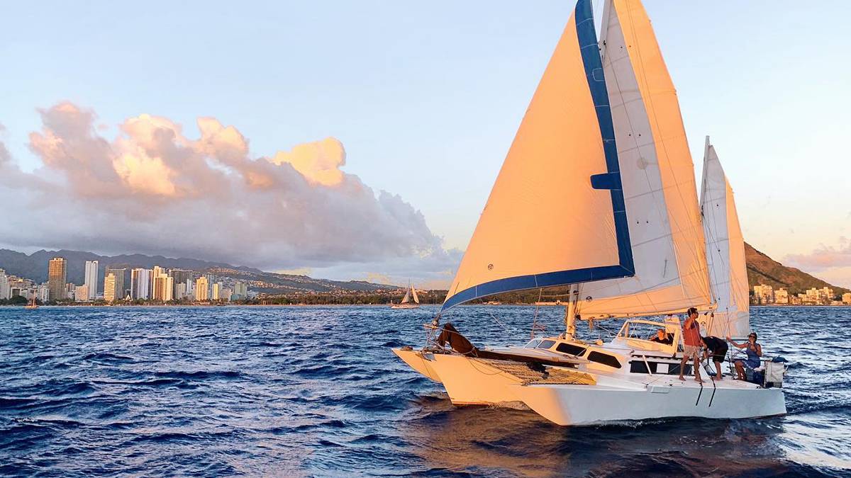 A white sail boat with white and blue sails and people on it on the water at sunset in Oahu, Hawaii