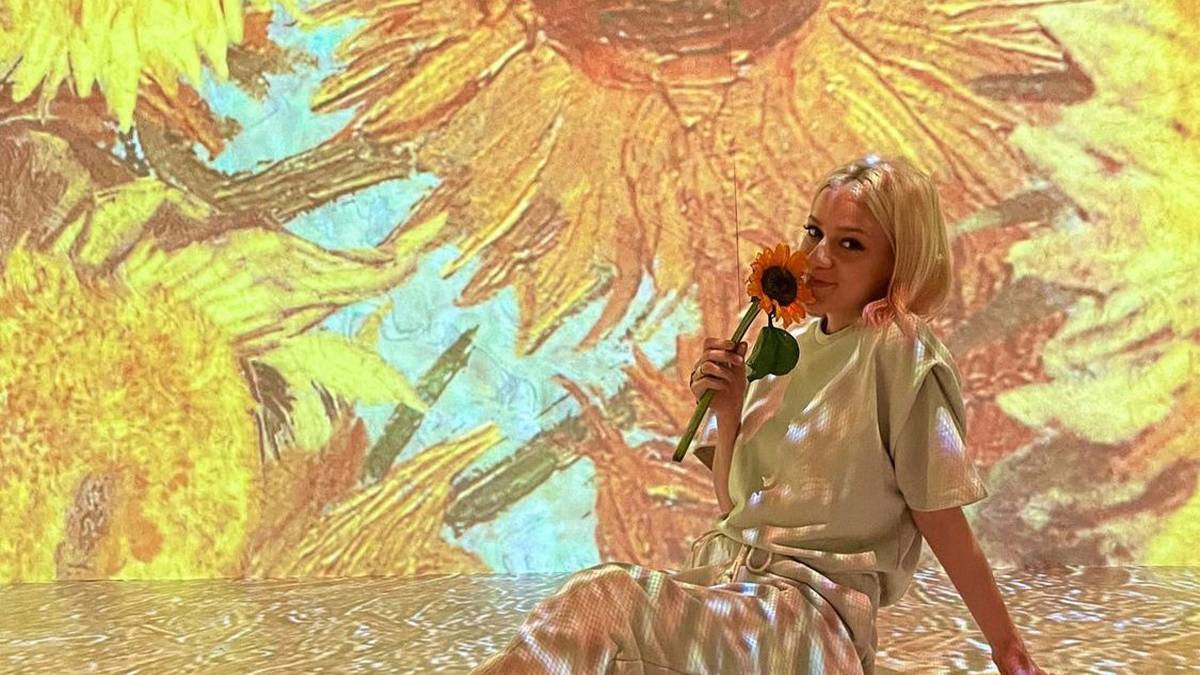 Young white woman holding a sunflower posing in front of a project of Van Gogh painting "Sun Flowers" in New York City, New York