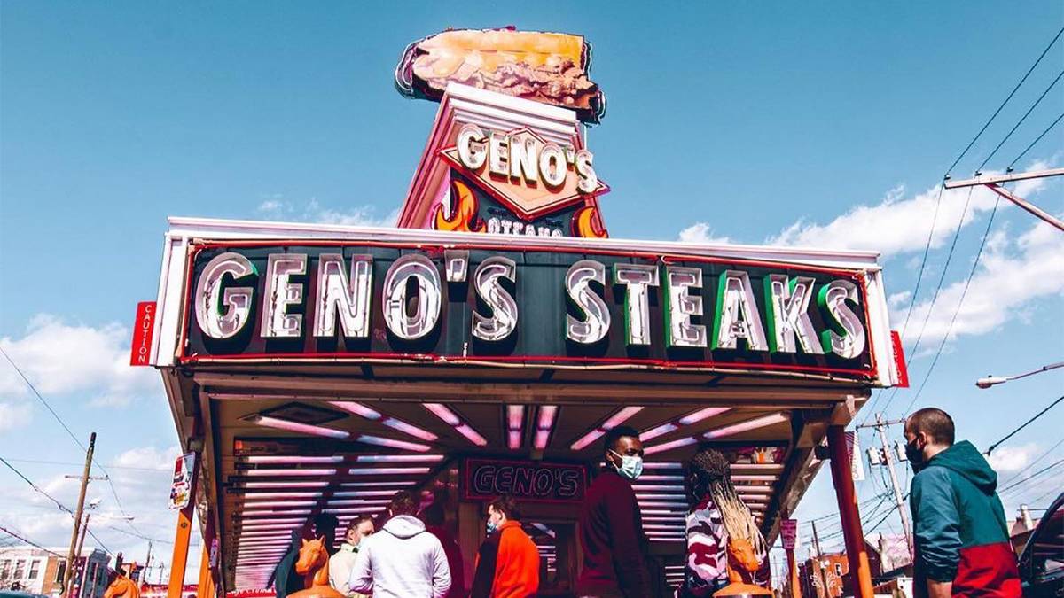 Looking up at a large Geno's Steak sign on the front of the restaurant with people waiting to order and a blue cloudy sky in the background in Philadelphia, Pennsylvania