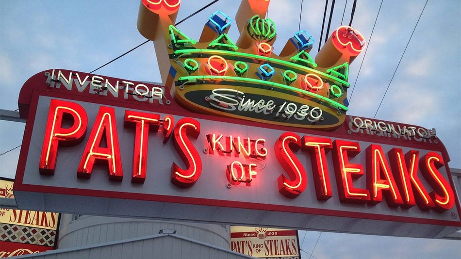 Looking up at a bright neon sign that says "Pat's King of Steaks" with a neon crown on top of it in Philadelphia, Pennsylvania