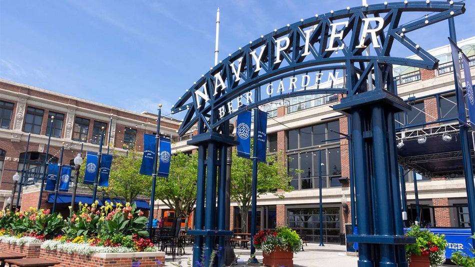 The blue iron entrance with brick planters and flower pots and white letters saying Navy Pier Beer Garden on a sunny day with building in the back ground in Chicago, Illinois, USA
