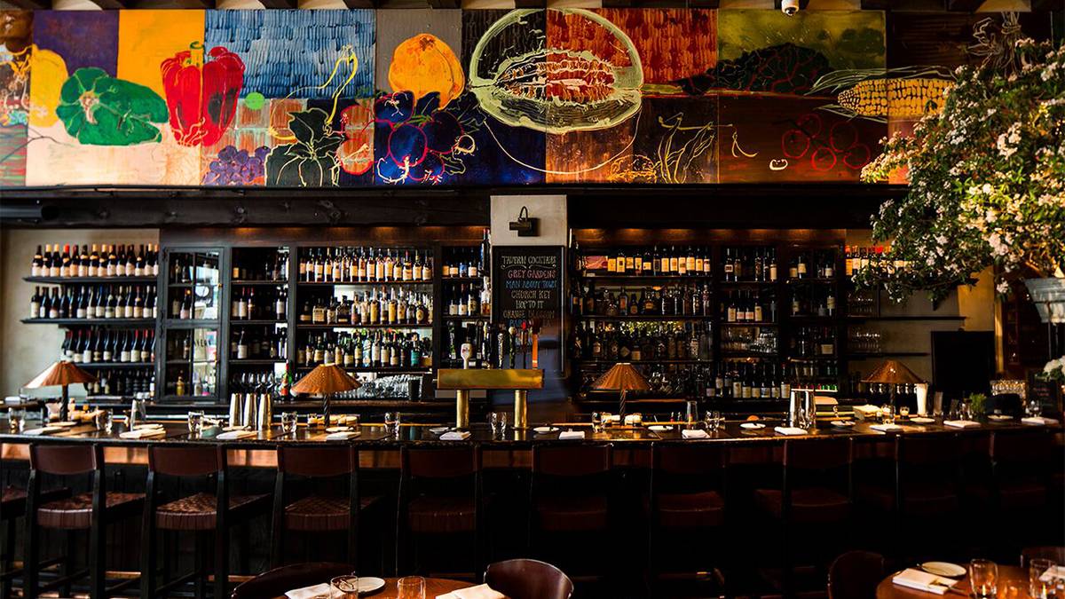 Straight on view of the bar lined with chairs and the wall behind the bar filled with bottle with a painting of fruits and vegetables over head at Gramercy Tavern in NYC, New York, USA