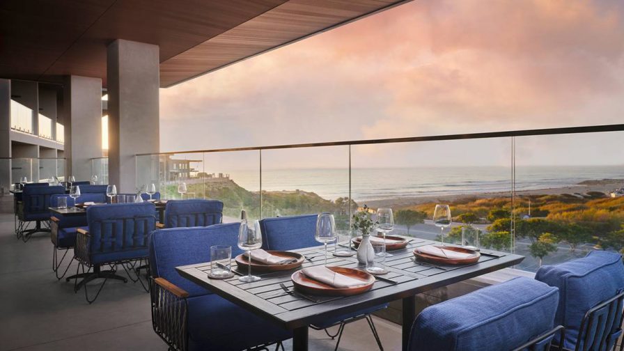Out door dining area with blue chairs and wooden tables with a pink sunset over the ocean behind it at VAGA Restaurant & Bar in San Diego, California, USA