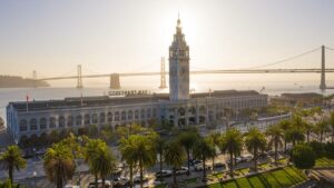 Aerial view of the Ferry Building at sunset with a row of palm trees in front of it and the Golden Gate Bridge behind it in San Francisco, California, USA
