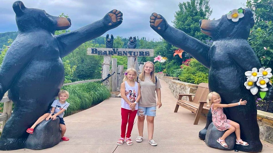 2 kids sitting on black bear statues' feet with two kids in the middle on a pathway at anakeesta with bearventures sign behind them surrounded by green mountains