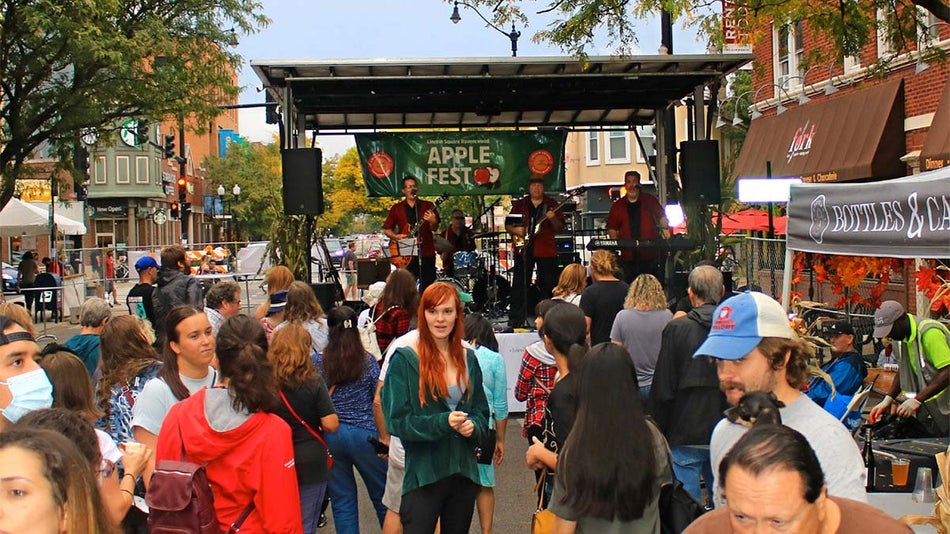 crowd of people gathered in front of stage with band playing set up on middle of street at Apple Fest with stall on the side in Chicago, Illinois, USA