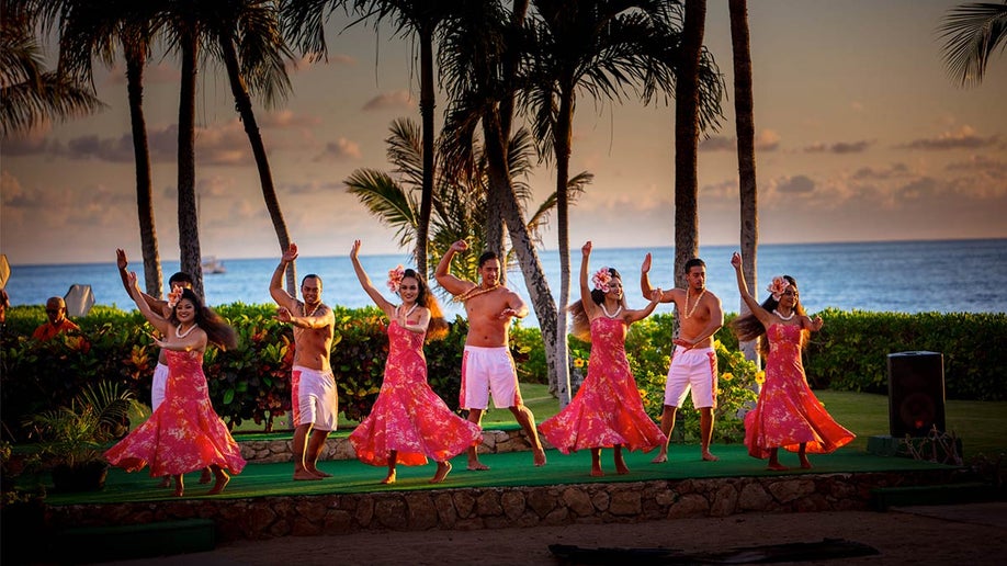 Hula dancers on stage with trees and view of water in background at Paradise Cove Luau in Oahu, Hawaii, USA