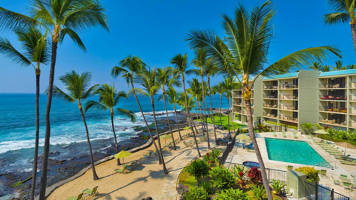 hotel with open balconies next to a resort style pool with palm trees and the ocean splashing onto black lava rock