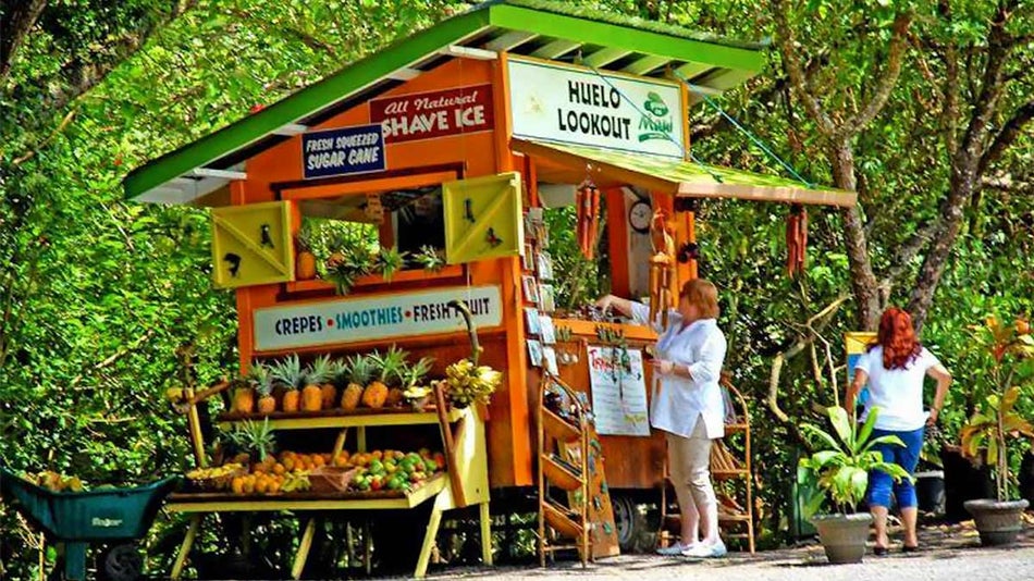 women in front of colorful stand decorated with various signs and filled with different fruits with trees in the background at Road to Hana in Maui, Hawaii, USA