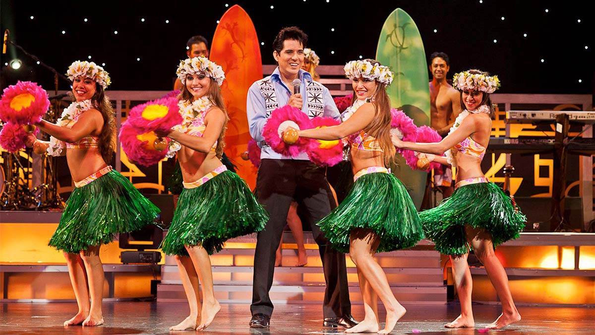 The man is singing while the women dance on stage and wear Hawaiian costumes.