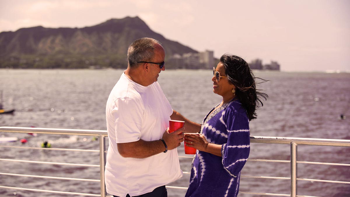 couple holding drinks at Spirit of Aloha Catamaran Sunset Sail with water in background and mountain and buildings in distance in Big Island, Hawaii, USA