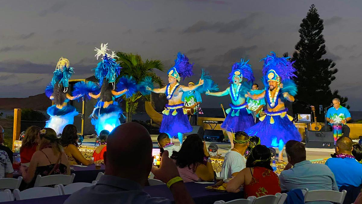 The people are watching the stage where a man is dancing in a blue Hawaiian attire 