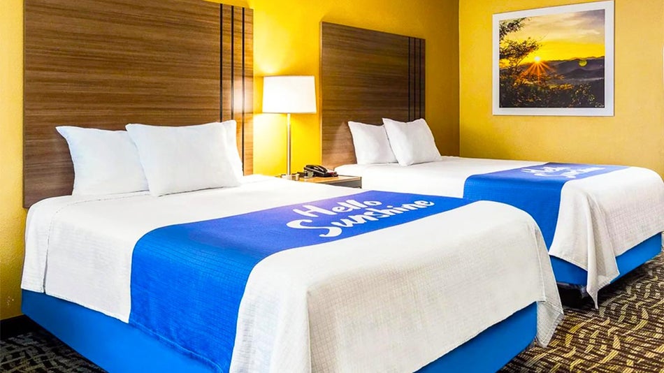 guest room with yellow walls and bed with white bed sheets with large blue stripe with words and wooden headboard at Days Inn Wyndham Buena Park in Los Angeles, California, USA