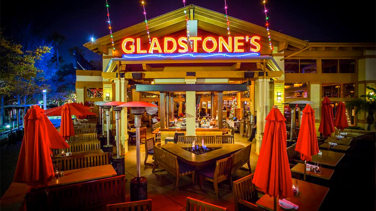 outdoor seating at Gladstones with dining tables and chairs and umbrellas and large neon sign on roof during night in Long Beach, California, USA