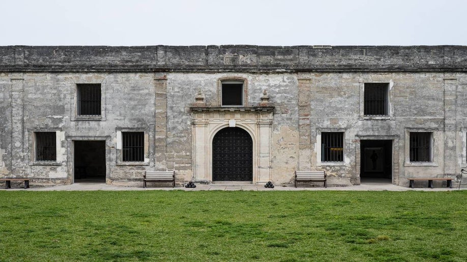 Stone walls of old fort with black doors and bars on window with green grass and grey sky