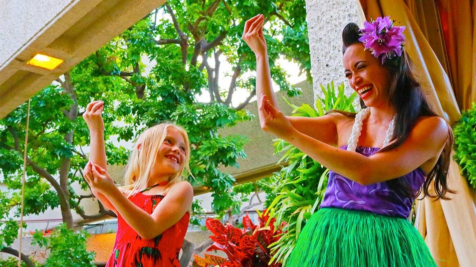 woman in grass skirt with flower in hair and child in red dress dancing with plants in background during day in Oahu, Hawaii, USA