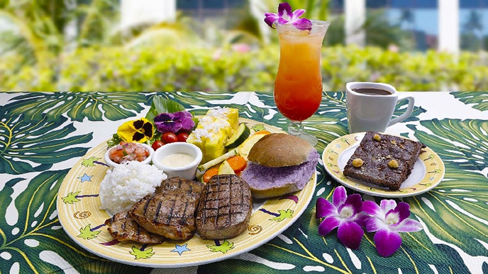 close up of plates of food with drinks on table with table cloth with pattern with plants in background during day at Waikiki Luau in Oahu, Hawaii, USA