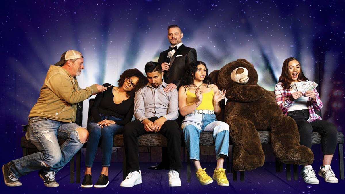 various people and a teddy bear sitting on a couch during a comedy hypnosis show with purple background