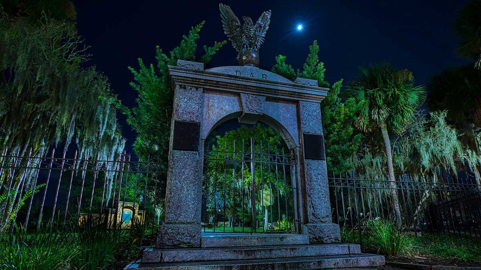 Stone entrance to a cemetery with an iron fence and trees at night