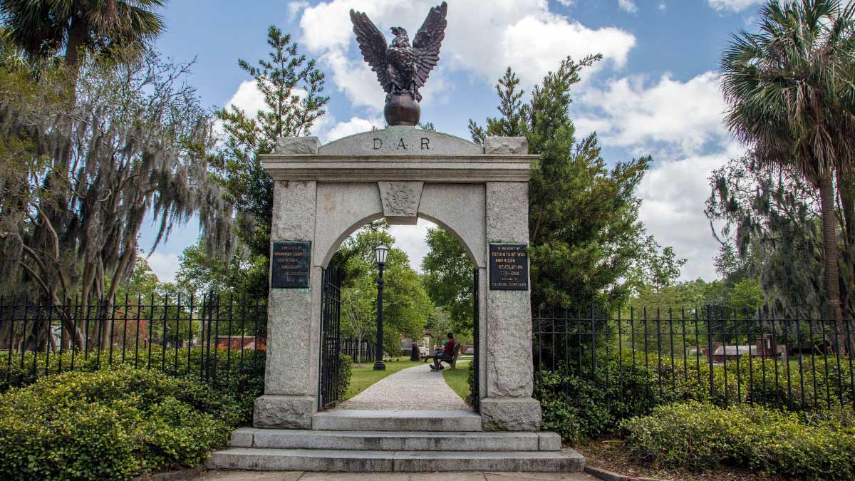 gated entrance to Colonial Park Cemetery with eagle statue on arch surrounded by greenery on a sunny day in Savannah, Georgia, USA