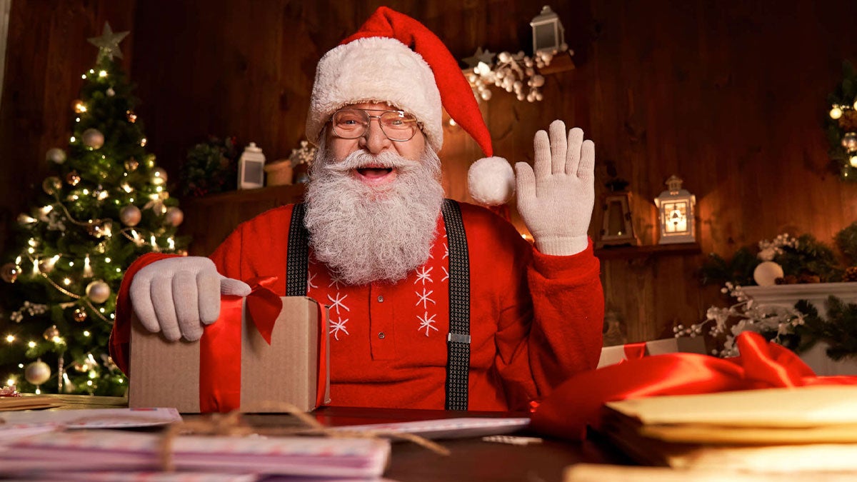 man dressed as santa on desk waving hand right hand with left hand resting on gift in room with christmas decorations and wooden walls at Cookies with Santa Zoo Atlanta in Atlanta, Georgia, USA