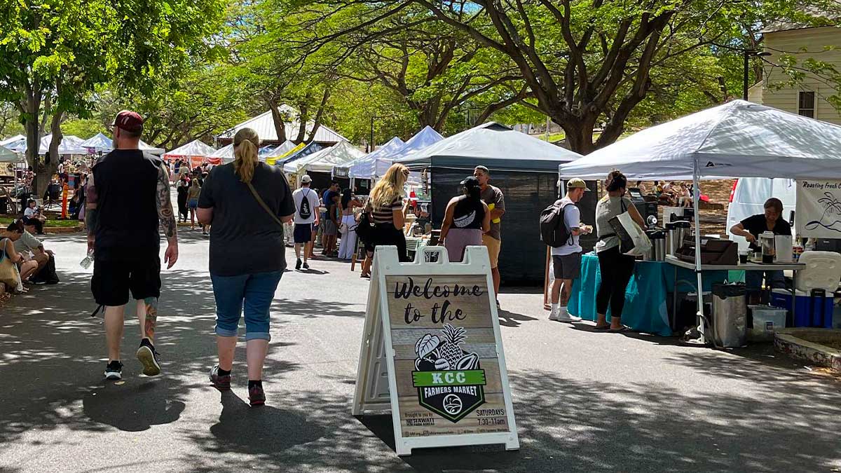people looking through stalls at KCC Farmers Market on a sunny day with welcome sign in the foreground in Oahu, Hawaii, USA