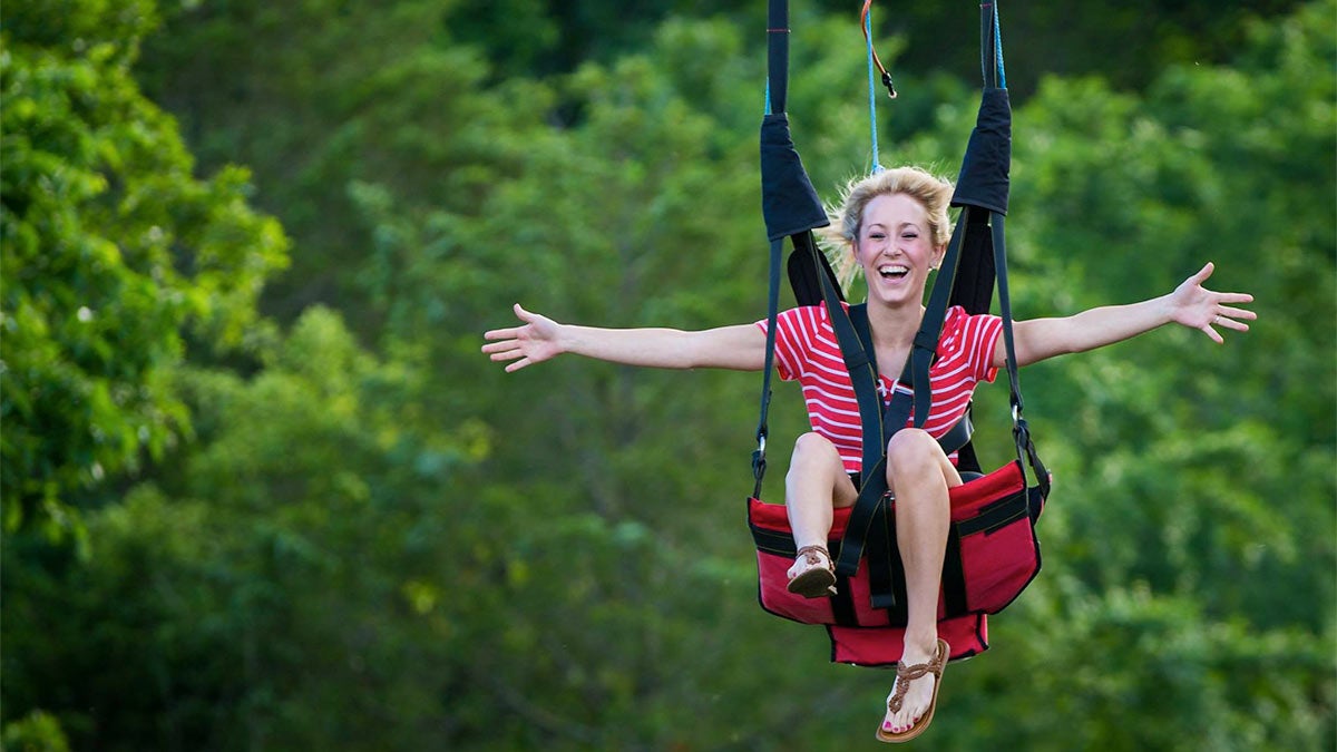 smiling woman in striped red and white shirt with arms extended riding down zipline surrounded by greenery on sunny day at Shepherd of the Hills Vigilante ZipRider in Branson, Missouri, USA