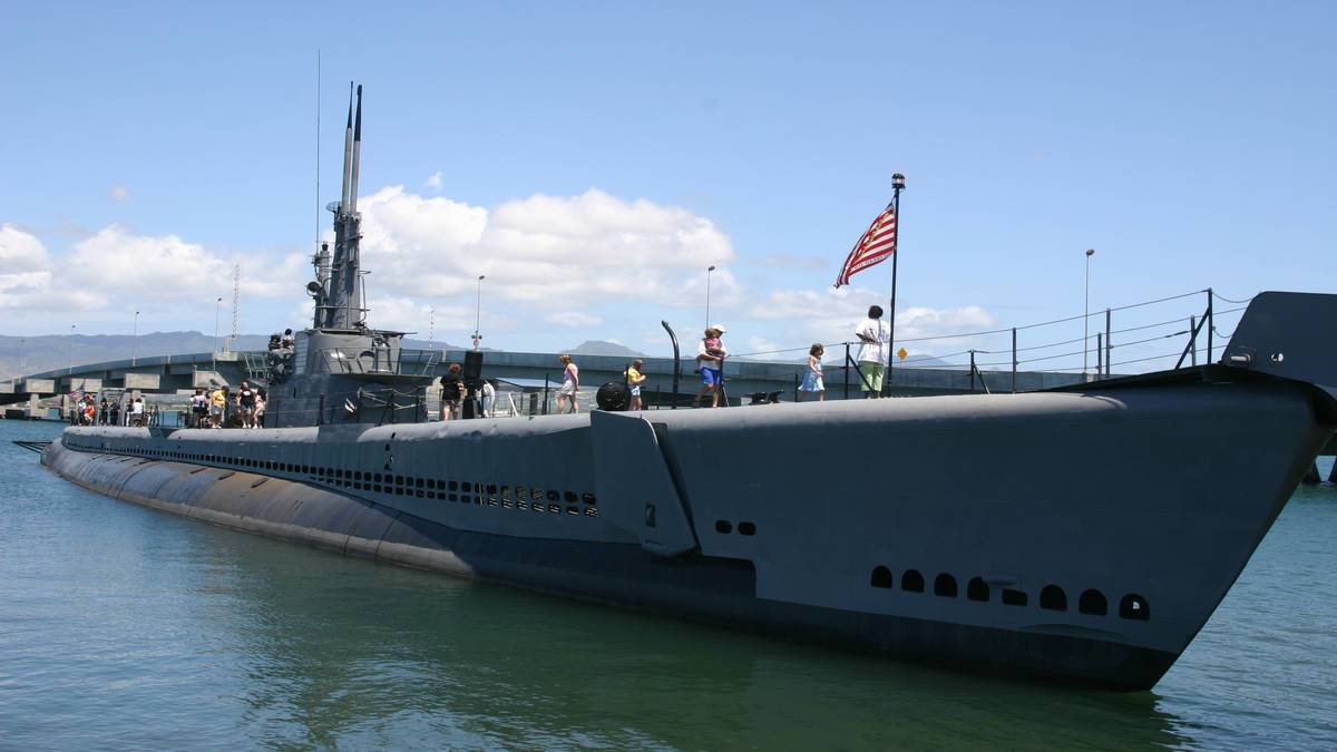 wide shot of the USS Bowfin in a harbor with people strolling on top and an American Flag blowing in the wind under a blue sky 