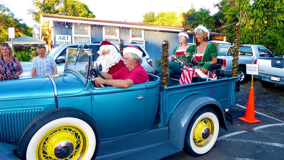 An old blue short bed pickup truck with Santa and Elves in it driving down the road at the Holualoa Village Association’s Annual Music & Light Festival in Maui, Hawaii, USA.