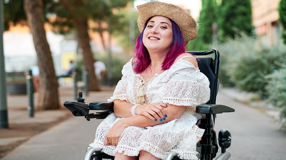 Woman smiling with purple hair wearing white dress and hat seated on wheelchair with trees and plants in background on a sunny day