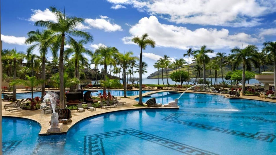 outdoor pool with palm trees, statues, and fountains surrounded by ocean and tropical foliage