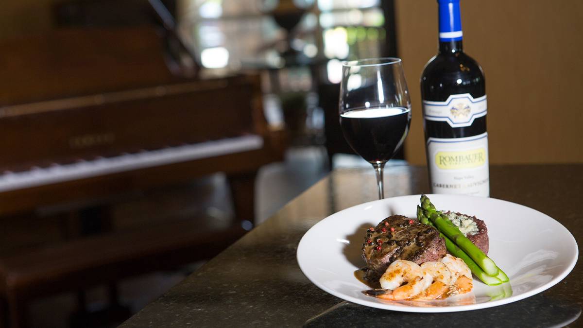 Close up photo of a surf and turf dinner, a glass of wine and a bottle next to it with a piano in the background at Chateau Grille in Branson, Missouri, USA