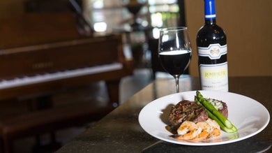 Close up photo of a surf and turf dinner, a glass of wine and a bottle next to it with a piano in the background at Chateau Grille in Branson, Missouri, USA