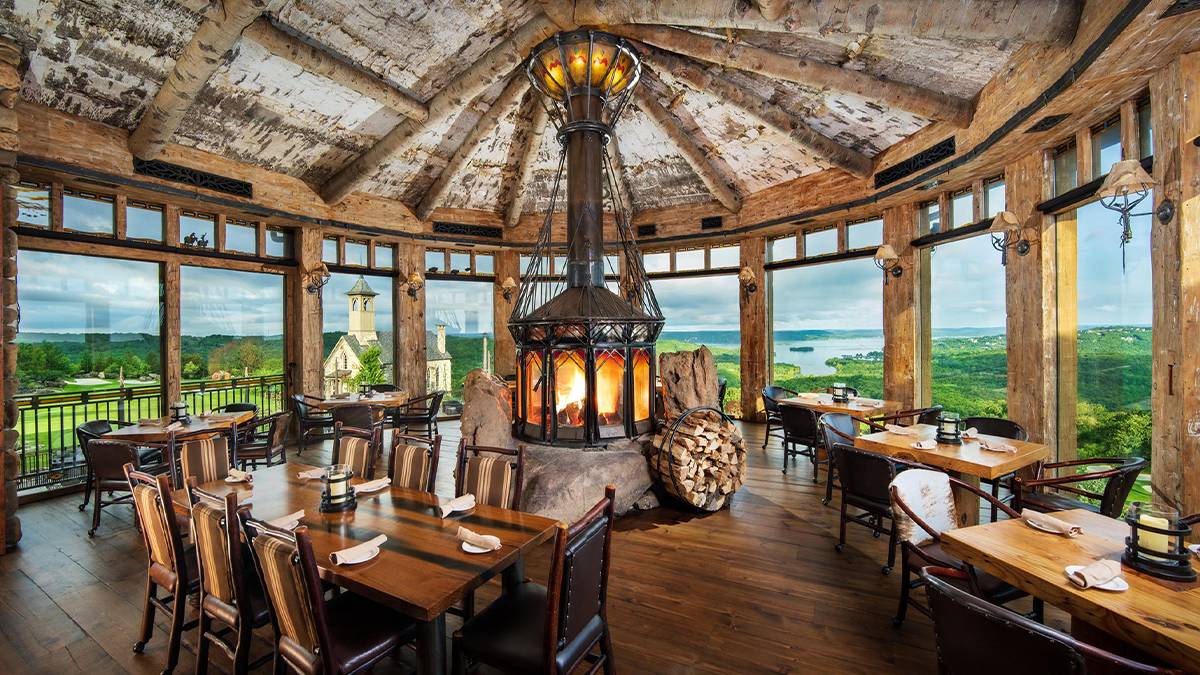 Wide shot of the dining room at Osage Restaurant at Top of the Rock, with an all the decor being wooden and a wood stove in the middle plus large windows that show a beautiful view of the Ozarks on a cloudy day in Branson, Missouri, USA