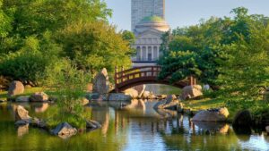 Wide shot of an arched bridge over water surrounded by large rocks and different types of trees and their reflections in the water and tall building in the back ground at the Osaka Japanese Garden in Chicago, Illinois, USA