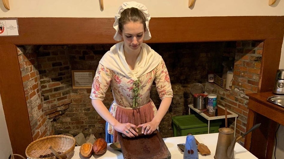 Woman in Colonial clothing making chocolate in front of a fireplace.