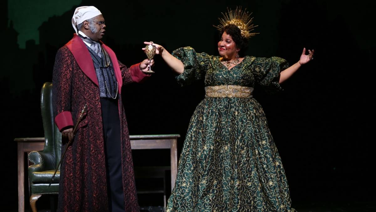 Two actors on stage, one with maroon robe and the other with gold and green dress holding a cup