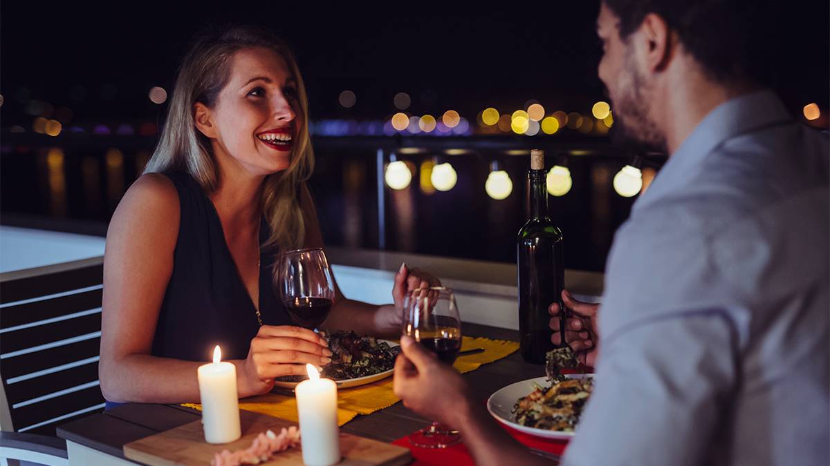 A couple having a romantic candle lit dinner outside at night with two glasses of red wine and lights in the background