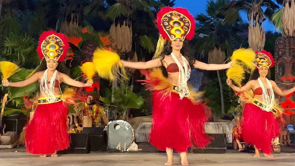 Three hula dancers in bright red and yellow costumes dancing on stage at Chiefs Luau in Honolulu, Hawaii, USA